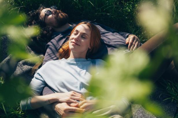 Zwei Personen liegen entspannt auf einer Decke im Gras, die eine Person trägt eine Brille.