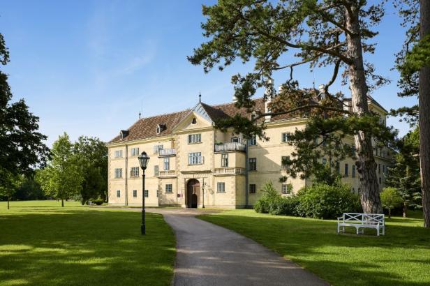 Schloss mit mehrstöckigem Hauptgebäude, Satteldach und verziertem Eingangsportal, umgeben von Rasenflächen, Bäumen und einem Weg mit Laterne und weißer Bank.