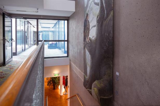 Blick von einem Treppenabsatz auf ein großes Wandgemälde eines Bären in einem modernen Gebäude mit Glasfenstern und Holzgeländer.