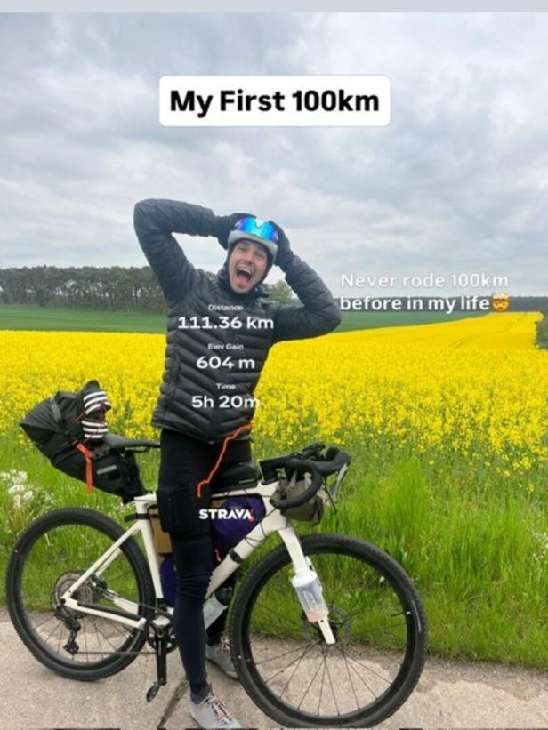 Ein Mann steht jubelnd mit Fahrrad vor einem gelben Rapsfeld, trägt Helm und Sportkleidung.