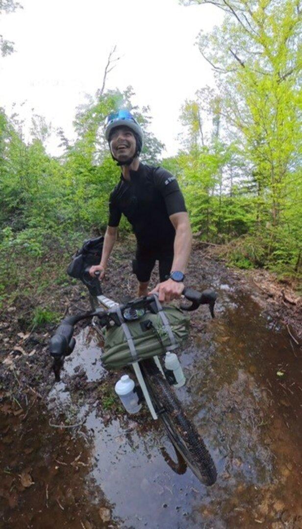 Ein Mann mit Fahrradhelm schiebt ein beladenes Fahrrad durch eine matschige Pfütze im grünen Wald.