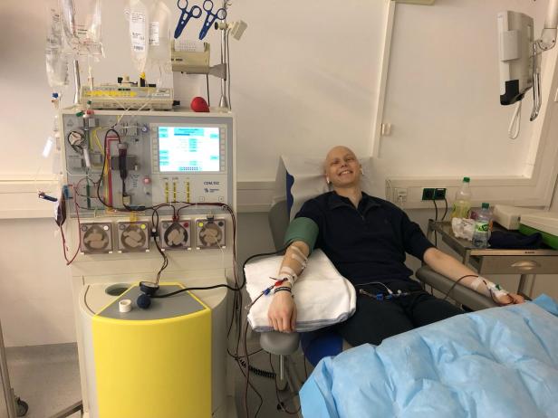 Eine Person sitzt lächelnd auf einem Stuhl und ist an eine medizinische Maschine zur Blutbehandlung angeschlossen.