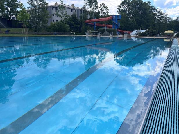 Leeres Schwimmbecken mit Schwimmbahnen im Freien, im Hintergrund eine rote Wasserrutsche und Bäume.