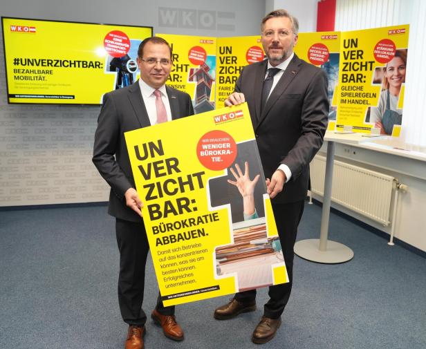 Zwei Männer im Anzug halten ein gelbes Plakat mit der Aufschrift „Unverzichtbar: Bürokratie abbauen“ in einem Büro.