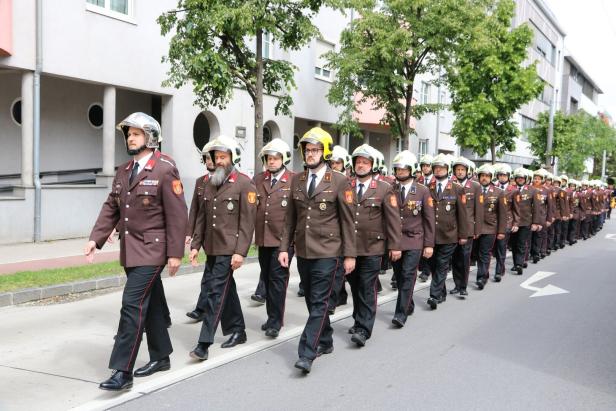Eine Gruppe uniformierter Feuerwehrleute mit Helmen marschiert in Reihen auf einer Straße entlang.