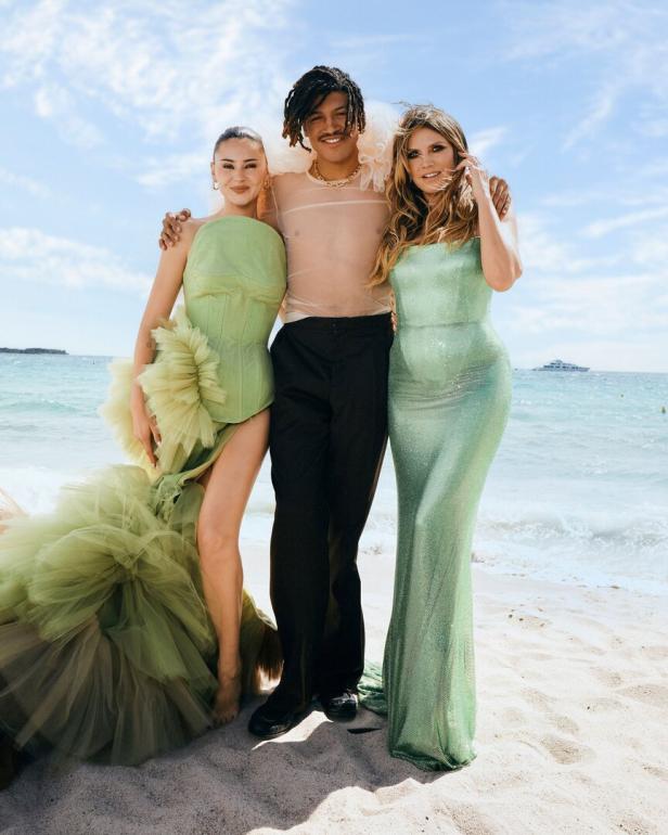 Drei Personen, darunter Heidi Klum, posieren am Strand vor blauem Meer und Himmel.