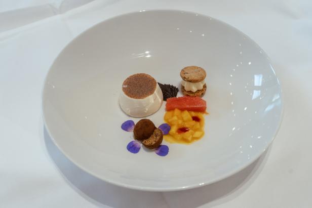 Ein kunstvoll angerichtetes Dessert mit Panna Cotta, Früchten, Pralinen, Keks-Sandwich und essbaren Blüten auf weißem Teller.