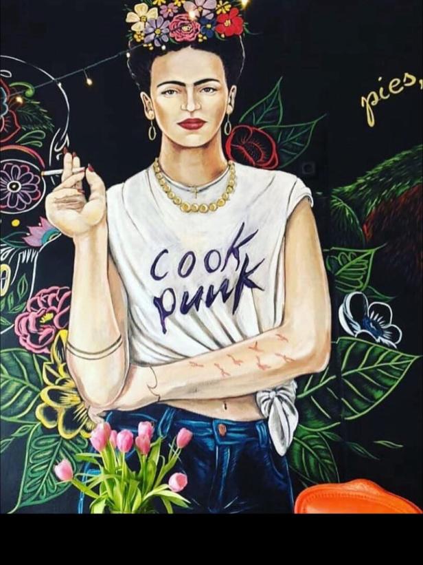 Eine Frau mit Blumenkranz, weißem Shirt mit „cook punk“, Zigarette in der Hand und bunten Blumen im Hintergrund.