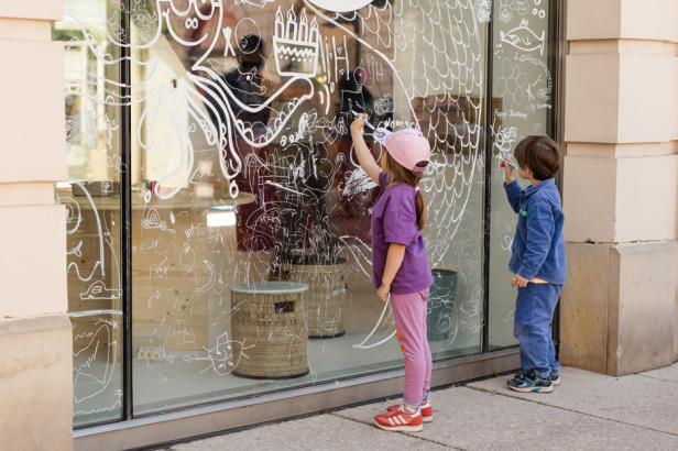 Zwei Kinder bemalen eine große Fensterscheibe mit weißen Zeichnungen und Motiven.