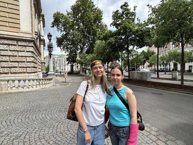 Zwei junge Frauen stehen auf einer gepflasterten Straße vor historischen Gebäuden und Bäumen.