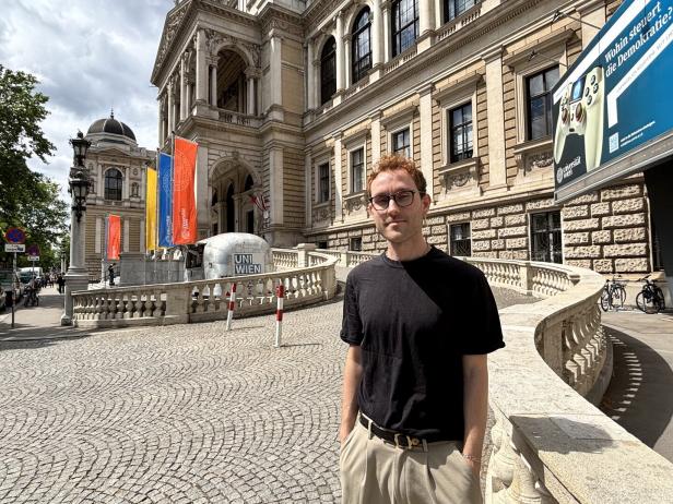 Ein Mann mit Brille steht vor einem historischen Gebäude mit Fahnen und der Aufschrift „UNI WIEN“ bei sonnigem Wetter.