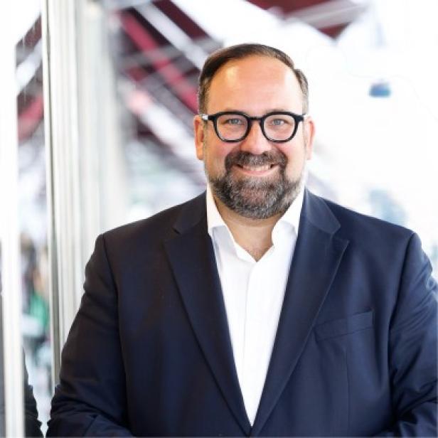 Ein Mann mit Bart und Brille trägt einen dunklen Anzug und steht lächelnd vor einer Glaswand.