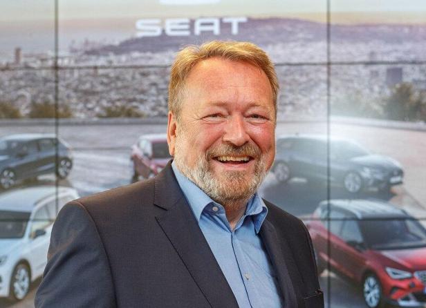 Ein lächelnder Mann im Anzug steht vor einer Wand mit dem Schriftzug „SEAT“ und mehreren Autos im Hintergrund.