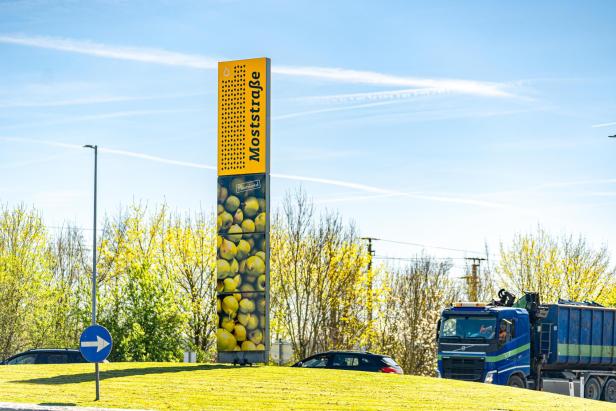 Gelbes Moststraße-Portal mit Birnenmotiv an einer Straße vor blühenden Bäumen und Fahrzeugen.