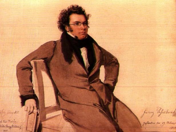 Franz Schubert sitzt lässig auf einem Stuhl, mit Brille und dunklem Mantel.