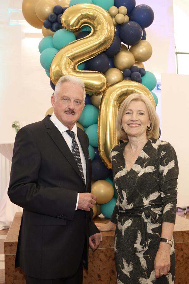 Ein Mann im Anzug und eine Frau im gemusterten Kleid stehen vor goldenen Ballons in Form der Zahl 20.