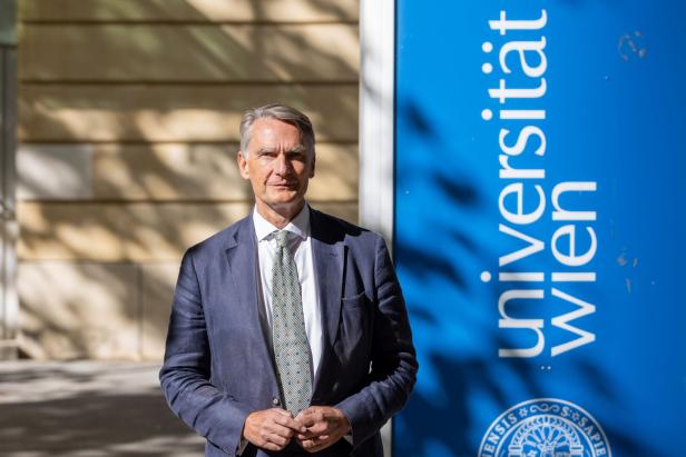 Ein Mann im Anzug steht vor einem blauen Schild der Universität Wien.