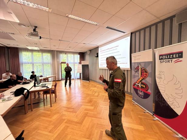 Ein Mann in Uniform hält einen Vortrag vor mehreren Zuhörern in einem Konferenzraum mit Präsentationsfolien und Polizei-Sport-Bannern.