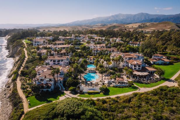 Weiße Gebäude mit roten Dächern, Palmen, zwei Pools und gepflegten Gärten direkt an einer Steilküste mit Meerblick.