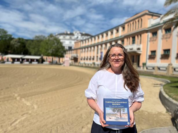 Eine Frau mit langen Haaren hält ein Buch vor einem historischen Gebäude mit sandigem Vorplatz und blauem Himmel.