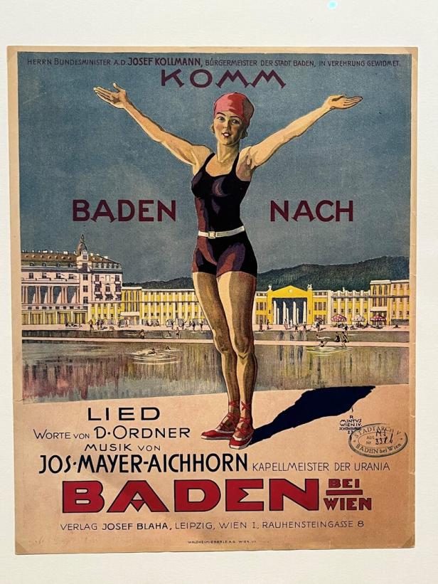 Eine Frau im Badeanzug und roter Badekappe steht mit ausgebreiteten Armen vor einem historischen Gebäude und Wasser.