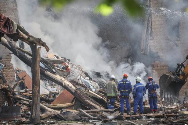 Vier Rettungskräfte stehen vor Trümmern und Rauch eines zerstörten Gebäudes.