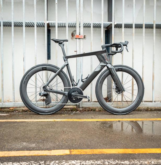 Ein modernes Rennrad mit aerodynamischem Rahmen steht vor einem Metallzaun auf nassem Asphalt.