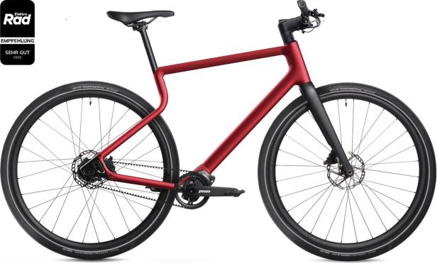 Rotes E-Bike mit schwarzer Gabel, Riemenantrieb, Scheibenbremsen und sportlichem Design, daneben ein Testsiegel mit Bewertung.