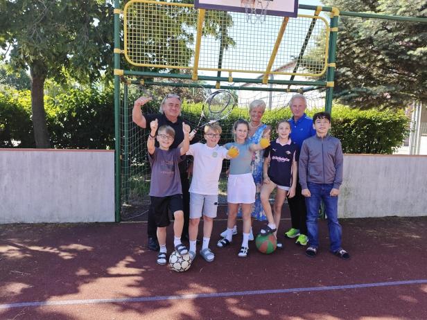 Sechs Kinder und drei Erwachsene stehen fröhlich auf einem Sportplatz vor einem Basketballkorb, einige halten Bälle in der Hand.