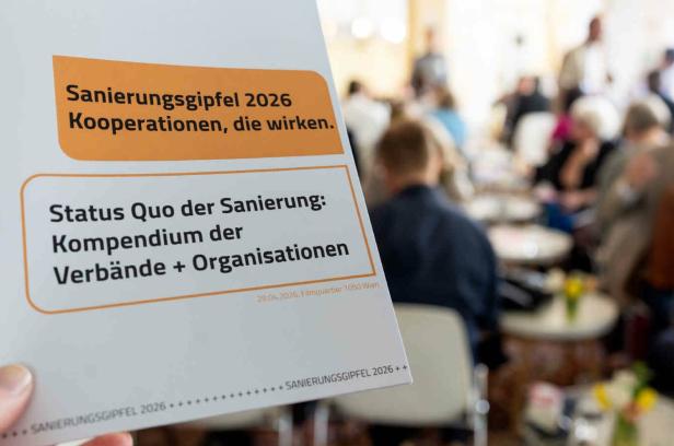 Hand hält Flyer mit der Aufschrift 'Sanierungsgipfel 2026 Kooperationen, die wirken. Status Quo der Sanierung: Kompendium der Verbände + Organisationen' vor unscharfem Publikum in einem Veranstaltungsraum.