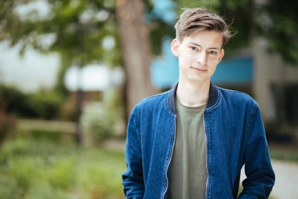 Junger Mann mit braunen Haaren trägt eine blaue Jacke und steht draußen vor unscharfem, grünem Hintergrund.
