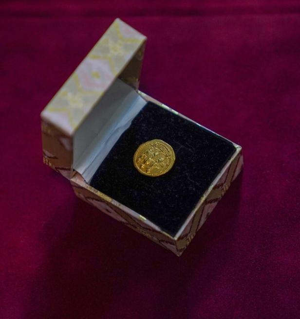 Eine goldene Münze liegt in einer kleinen, offenen Schachtel mit schwarzem Samt auf rotem Untergrund.