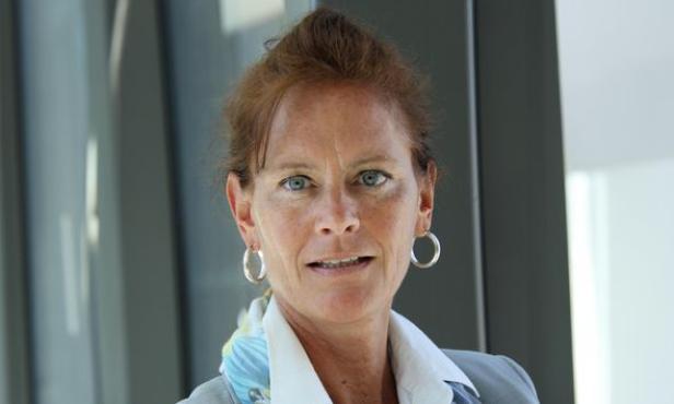 Frau mit blauen Augen, hellblauer Bluse und Ohrringen vor Glasfassade.