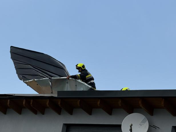 Ein Feuerwehrmann mit Helm repariert ein abgerissenes Blechdach auf einem Hausdach bei klarem Himmel.