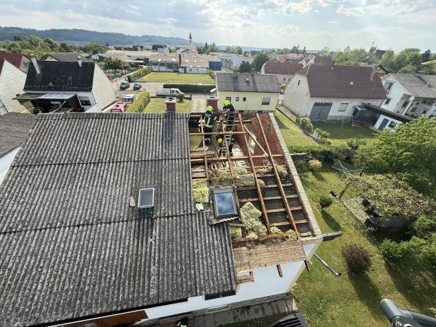 Mehrere Personen arbeiten auf einem teilweise abgedeckten Hausdach, umgeben von Wohnhäusern und Gärten.