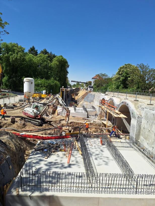Mehrere Bauarbeiter und Baufahrzeuge arbeiten an einer großen Baustelle mit Betonfundamenten und Stahlbewehrung unter blauem Himmel.