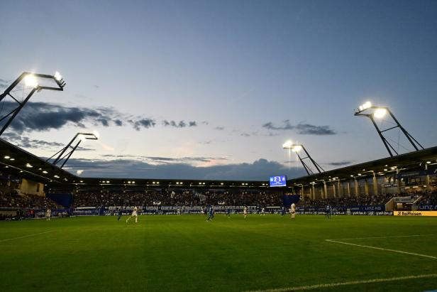 Ein beleuchtetes Fußballstadion bei Abenddämmerung mit Zuschauern auf den Tribünen und Spielern auf dem Spielfeld.