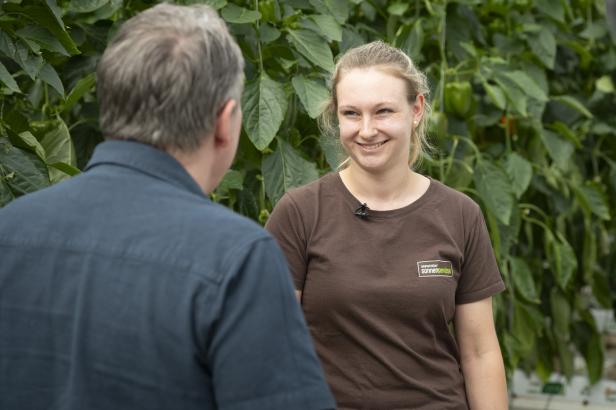 Zwei Personen im Gespräch vor grünem Pflanzenhintergrund, eine trägt ein braunes T-Shirt mit einem kleinen Logo auf der Brust.