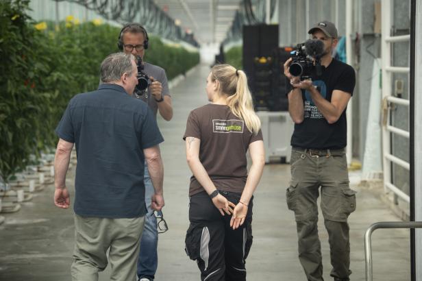 Zwei Personen gehen in einem Gewächshaus, zwei weitere filmen sie mit Kameras.