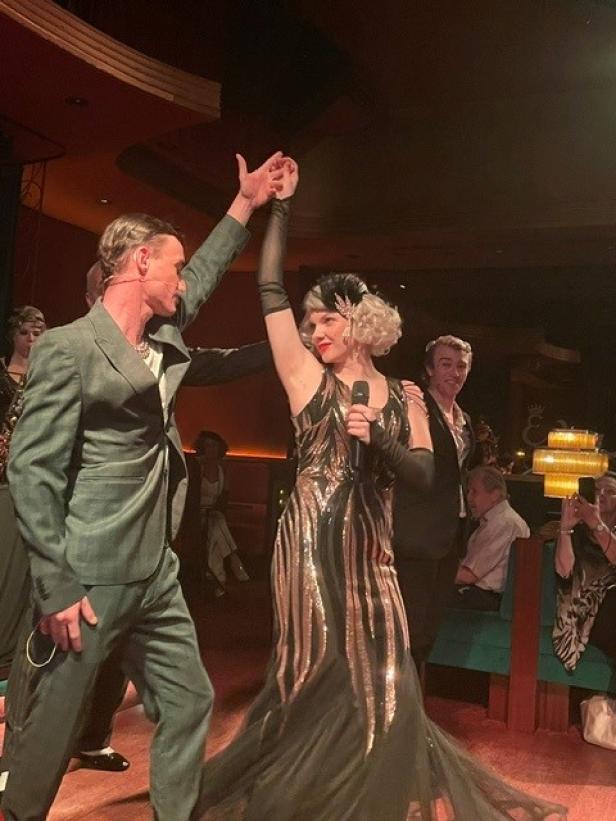 Schauspielerin im glamourösen Kleid mit Mikrofon auf Bühne, umgeben von weiteren Darstellern.