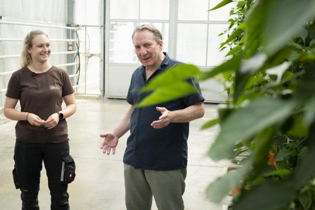 Zwei Personen stehen in einem hellen, industriellen Raum, eine trägt ein dunkles T-Shirt und Arbeitshose, die andere ein dunkles Hemd und helle Hose, im Vordergrund unscharfe grüne Blätter.