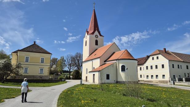 Eine Person geht auf eine Kirche mit rotem Turm zu, umgeben von gelben Gebäuden und Wiesen.