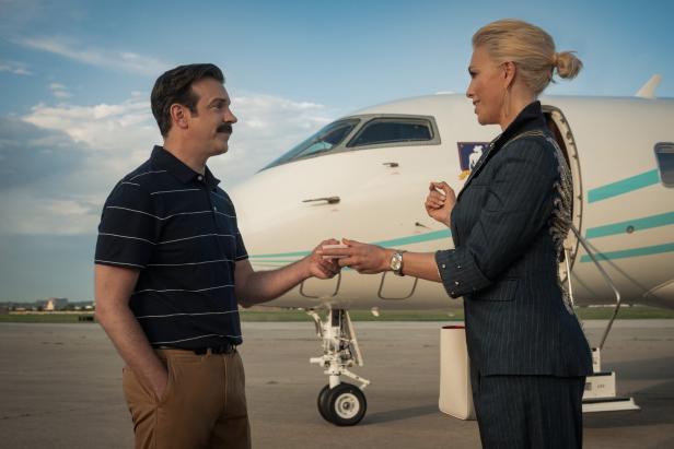 Ein Mann und eine Frau stehen vor einem Privatjet und reichen sich gegenseitig einen kleinen Gegenstand.