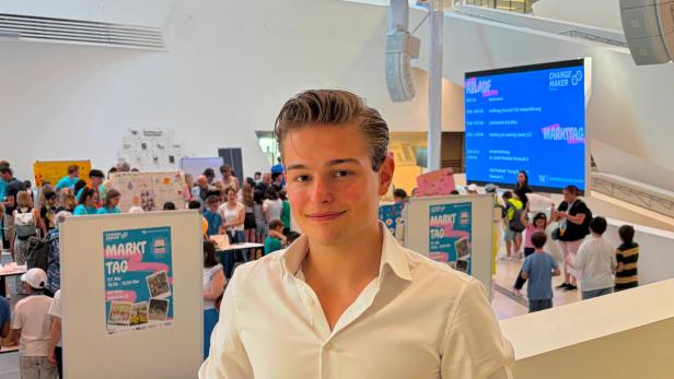 Junger Mann in weißem Hemd vor belebtem Markttag mit Kindern und Infoständen in heller Halle.