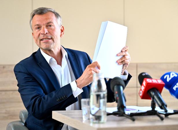 Ein Mann im Anzug präsentiert Dokumente vor Mikrofonen von ORF und anderen Sendern.