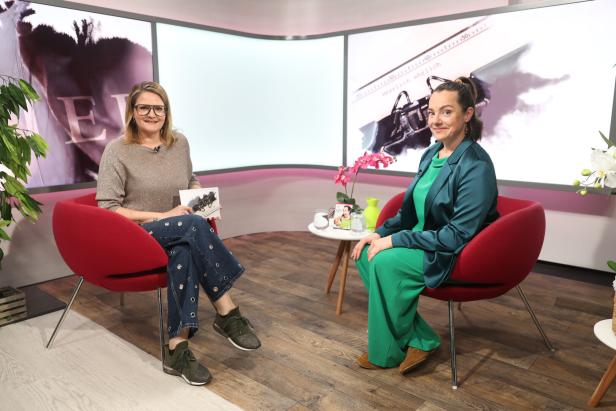 Zwei Frauen sitzen in roten Sesseln in einem modernen Studio, zwischen ihnen ein kleiner Tisch mit Getränken und Blumen.