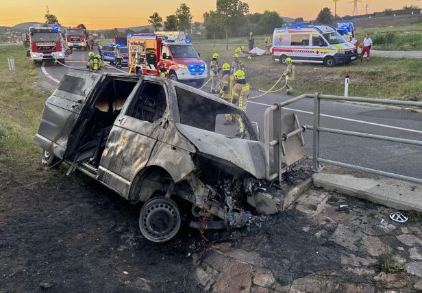 Ausgebrannter Transporter an Betonwand, Feuerwehr und Rettungskräfte im Einsatz.