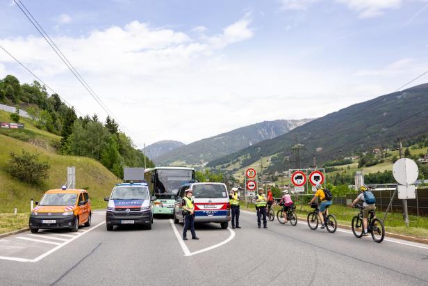 Mehrere Polizisten kontrollieren an einer Straßensperre Fahrzeuge und Radfahrer vor einer Berglandschaft.