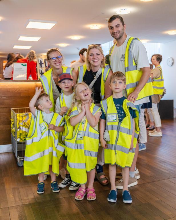Drei Erwachsene und vier Kinder tragen gelbe Warnwesten und lächeln gemeinsam in einem hellen Raum.