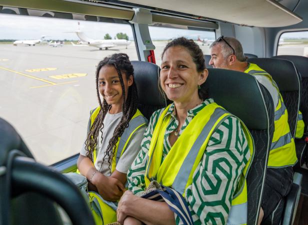 Zwei Personen mit Warnwesten sitzen lächelnd in einem Bus am Flughafen, im Hintergrund sind Flugzeuge zu sehen.